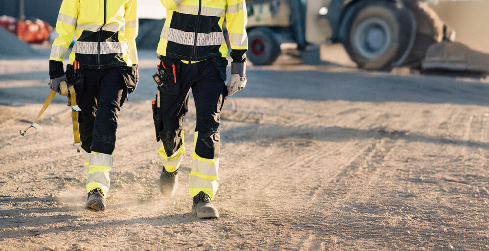 Två personer i varselbyxor och skyddsskor går på en grusplan